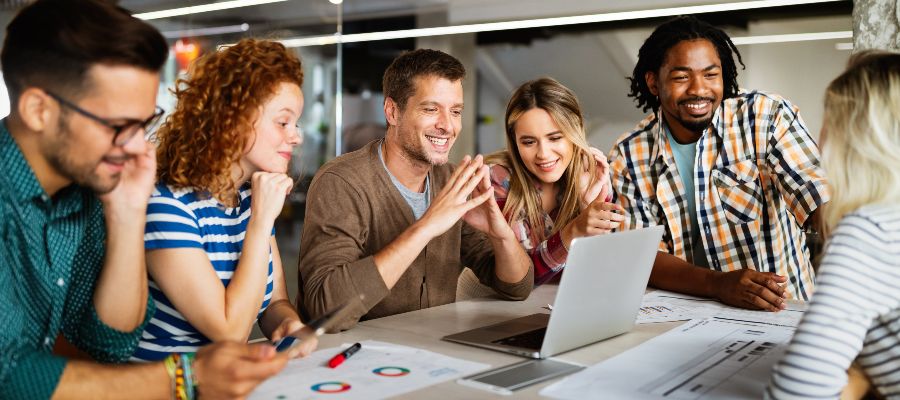 A group of smiling young people are standing around a table on which there is a laptop and some documents.