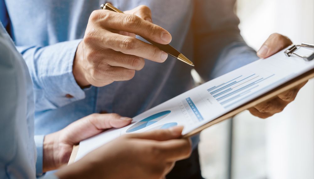 A close-up of two pairs of hands holding a clipboard with a diagram on it. One hand is pointing at the diagram with a pen.