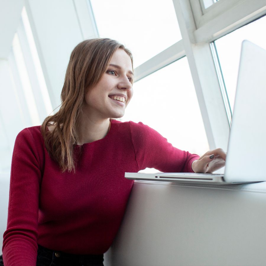 Une femme vêtue d'un pull rouge est assise devant une fenêtre, devant son ordinateur portable.